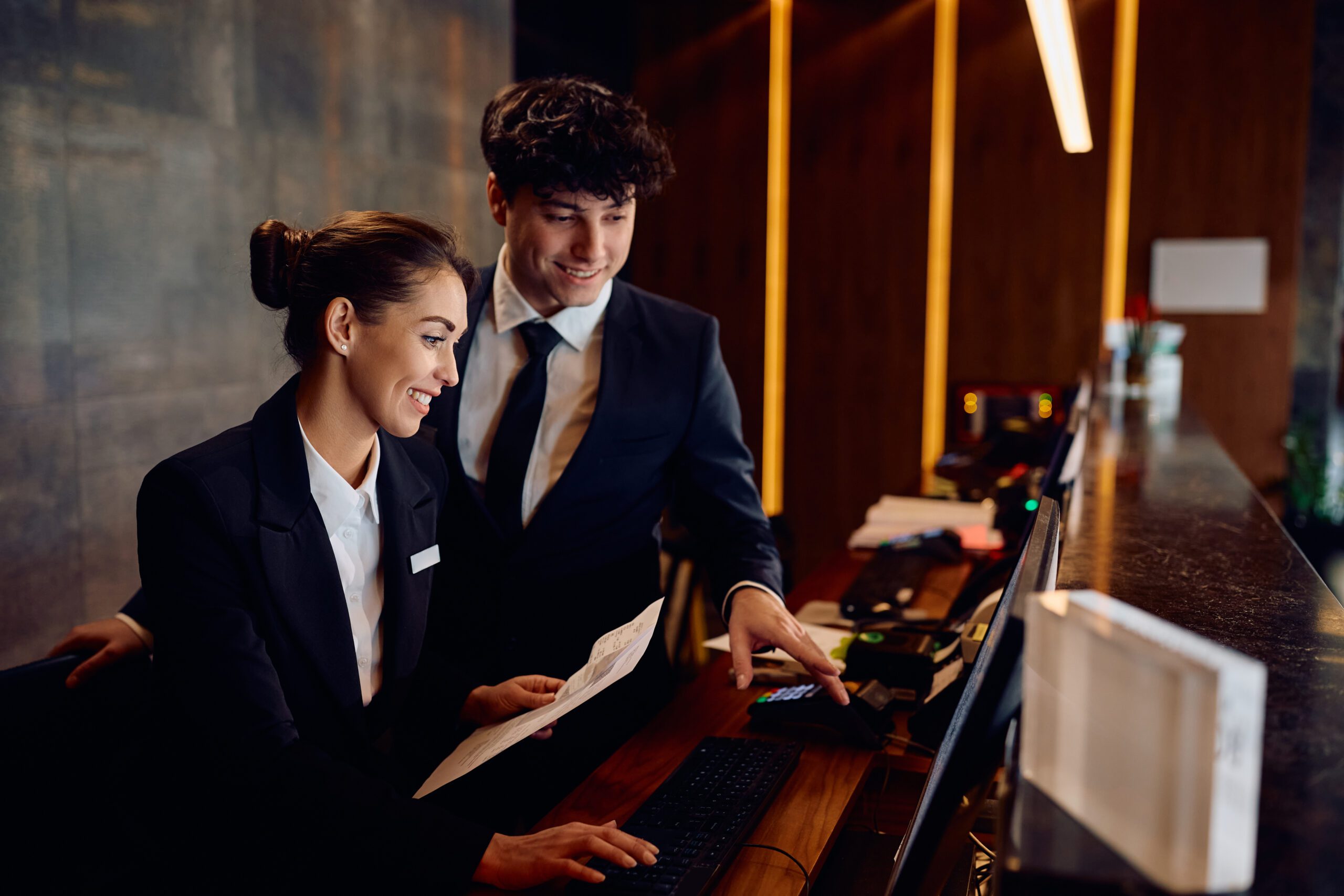 Happy receptionists cooperating while working on computer at hotel front desk.